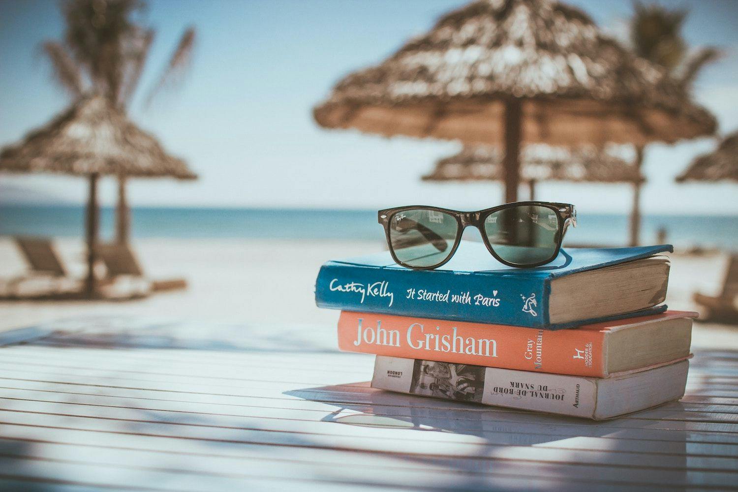 Image shows three books stacked on a table on a beach with a pair of sunglasses on top of them.