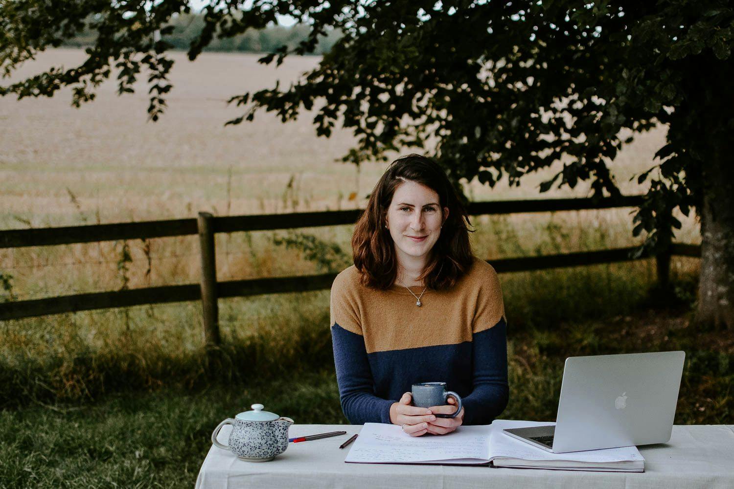 Josephine is sitting outside at a table with her laptop. She is holding a mug of tea and is smiling at the camera