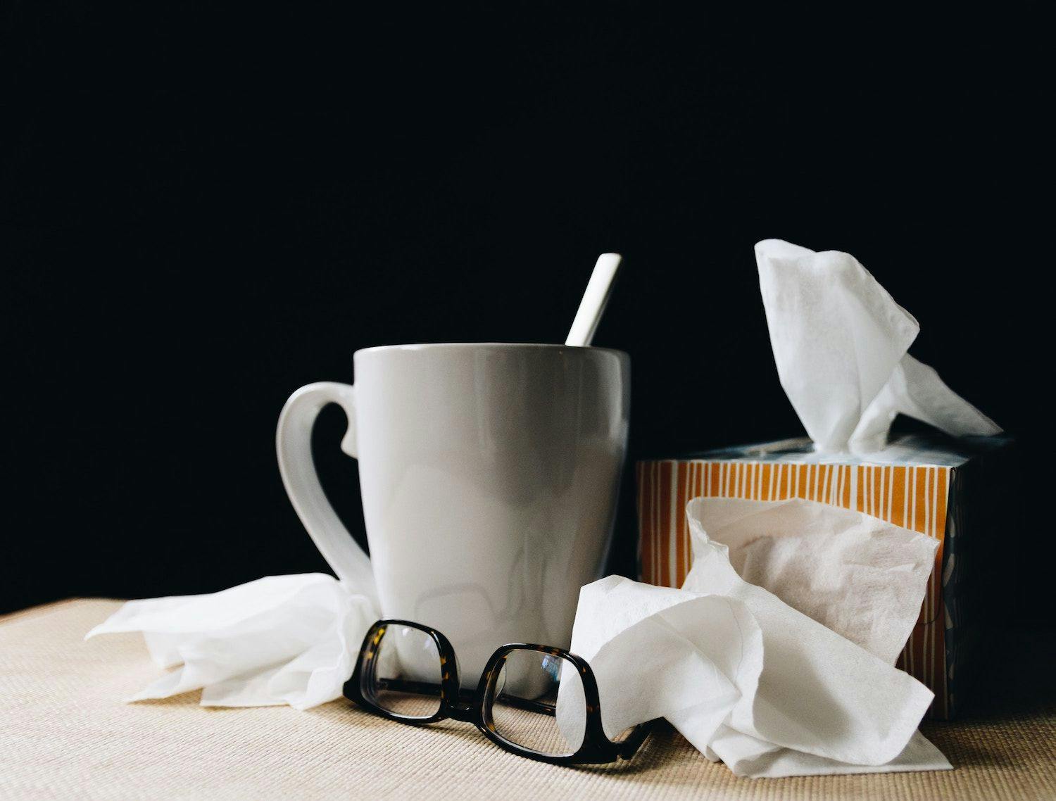 Image shows a mug of tea, a pair of glasses and a tissue box with tissues scattered.