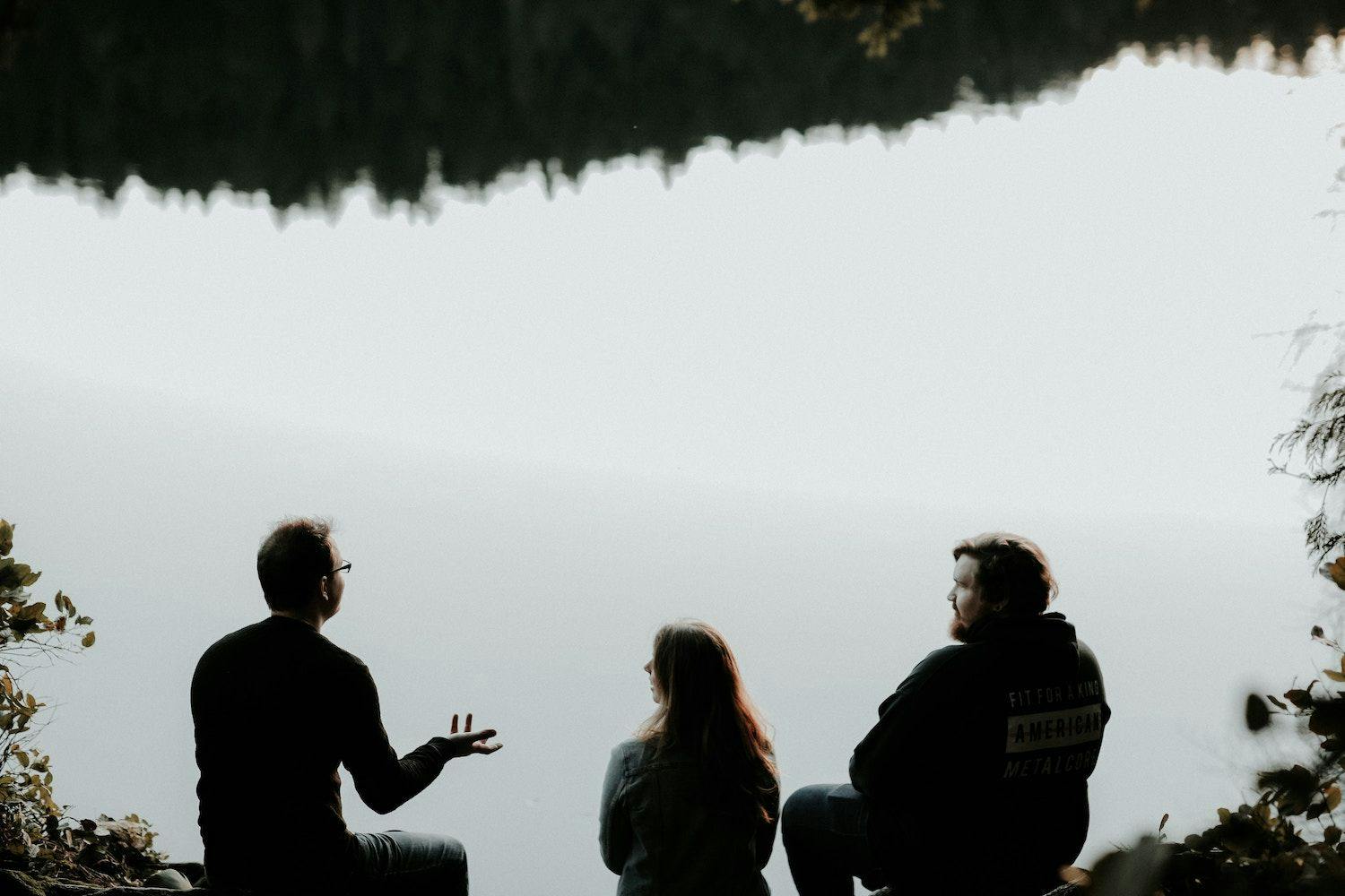 Three people sat by a lake talking.