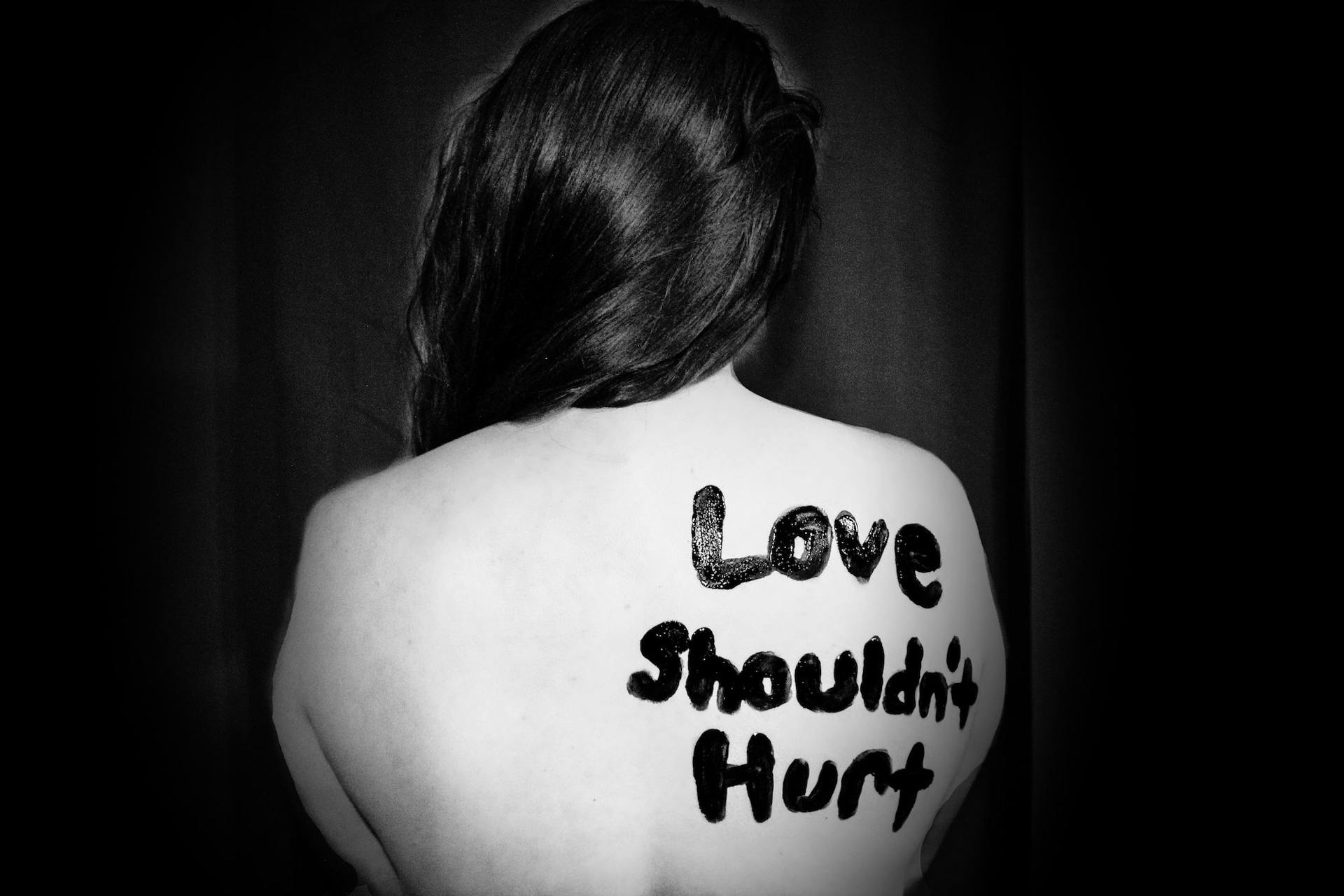Someone sits with their back to the camera, the words 'Love shouldn't hurt' written on their back in black paint