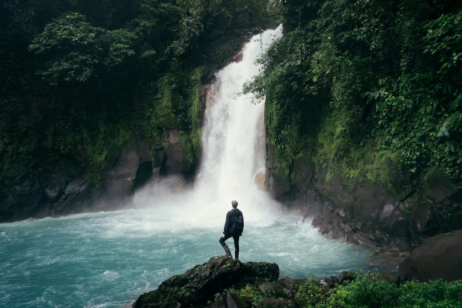 Image shows a person stood at the foot of a waterfall surrounded by greenery.