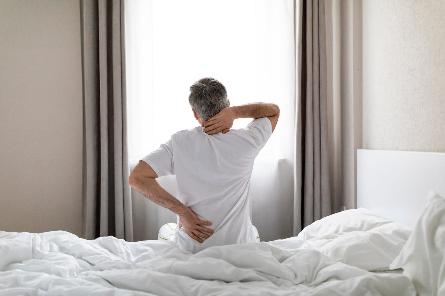 Image shows man getting out of bed holding lower back and neck.