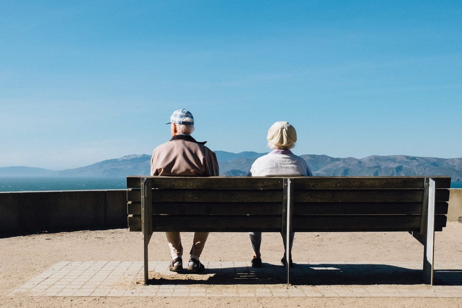 Image shows older couple sat on a bench admiring a sea view.